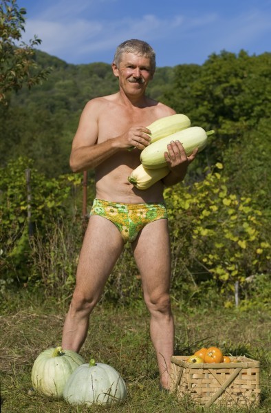 A man stands with vegetable marrows in his hands. At his feet are two pepos and box with tomatos.